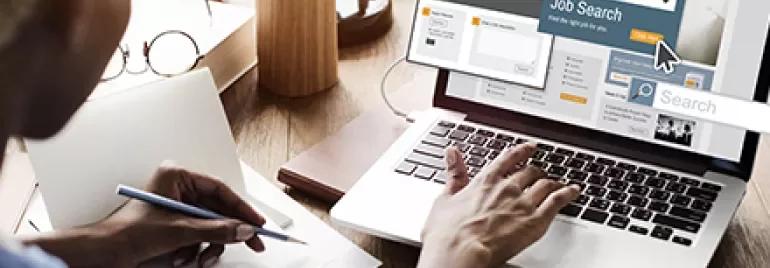 Person writing notes while using a laptop for job searching.