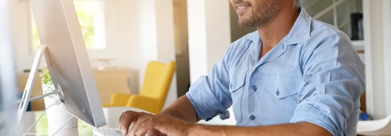 Man in glasses working at a desktop computer in a bright room.