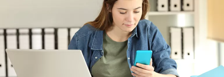Woman in denim shirt checks phone at desk with laptop.