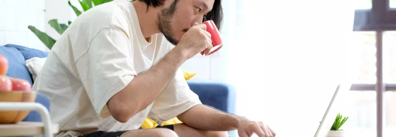 Man in a white shirt drinking coffee while working on a laptop.