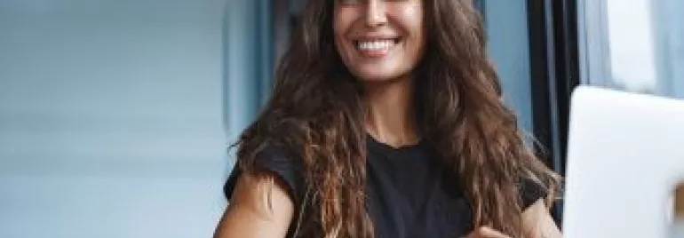 A woman with long hair smiles while working on a laptop by a window.