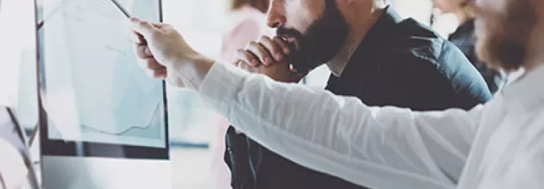 Two men working on desktop computers, discussing data on a screen in a modern office.