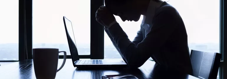 Silhouette of a person at a desk, head bowed near a laptop with a mug and papers.