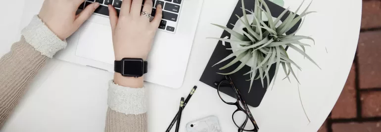 Hands typing on a laptop beside a plant and glasses on a white table.