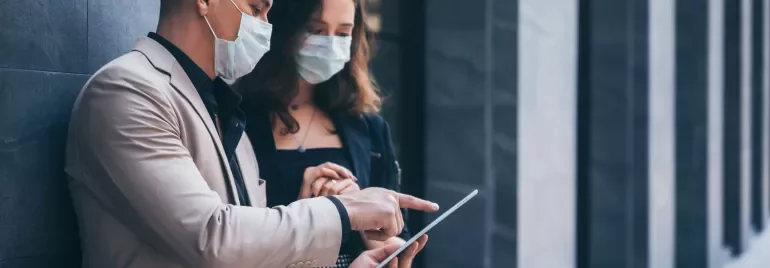 Two people in masks discussing on a tablet outdoors.