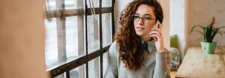 Woman with curly hair on phone, holding a mug, sitting by a window in a cozy cafe.