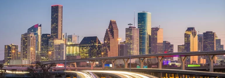 City skyline at dusk with illuminated skyscrapers and light trails on the highway.