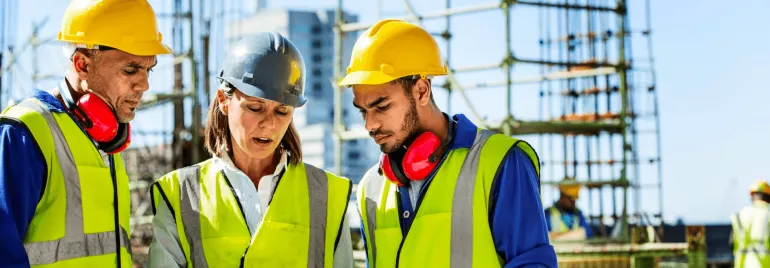 Three construction workers wearing safety gear discussing a project on site.