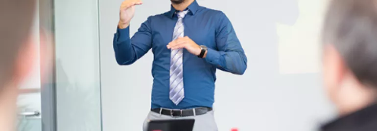Man giving a presentation in a blue shirt and tie, gesturing with hands.