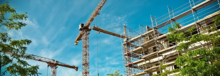 Three cranes towering over a building under construction with scaffolding against a blue sky.