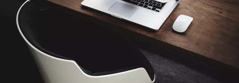 Open laptop and mouse on a wooden desk with a modern chair.