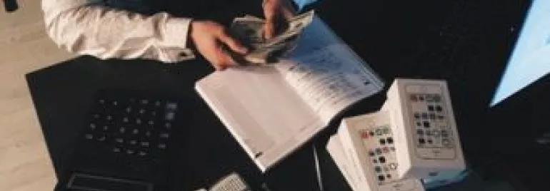 Person counting cash at a desk with electronic devices and a notebook.
