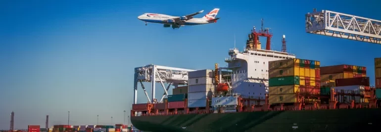 A cargo ship in port with colorful containers and an airplane flying overhead under a clear blue sky.