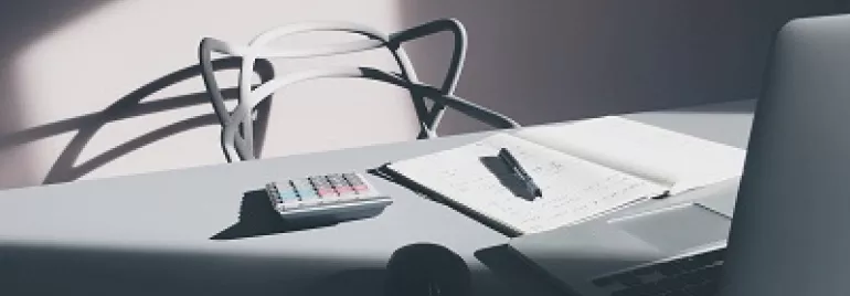 A laptop, notepad, pen, and calculator on a sunlit desk.