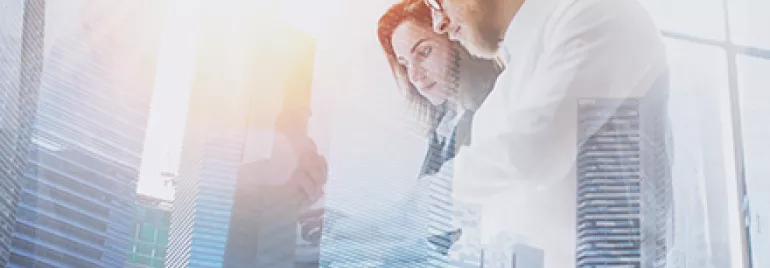 Double exposure of two business professionals working on a tablet with skyscrapers and sunlight in the background.