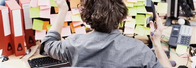 Person at desk surrounded by sticky notes and binders, holding a phone.