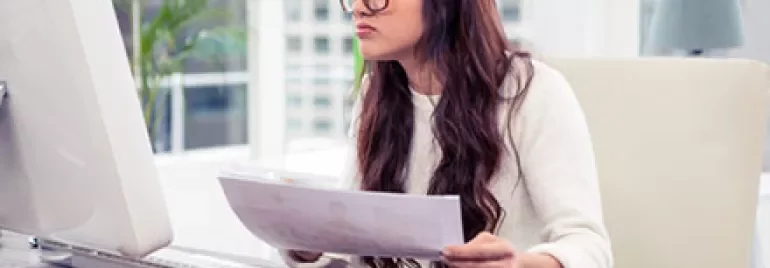 Woman in glasses working at a computer, holding documents.