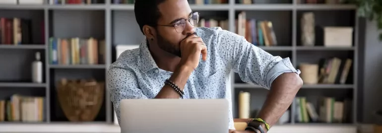 Man sitting at desk with laptop, thinking, bookshelf in background.