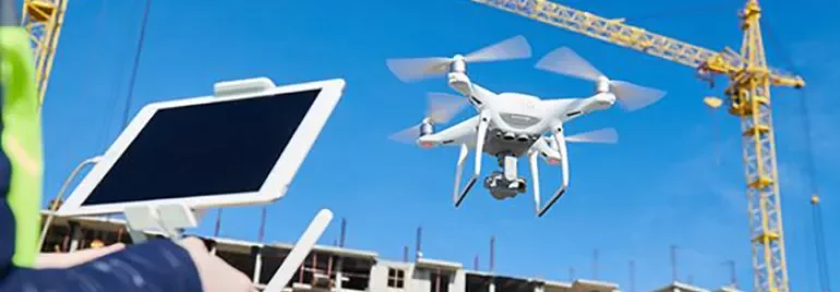 Person controlling drone with tablet at a construction site with cranes and building in the background.