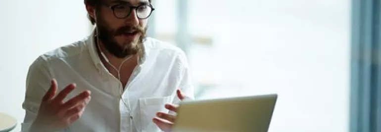Man wearing headphones, engaged in a video call on a laptop, sitting at a cafe with a cup of coffee.