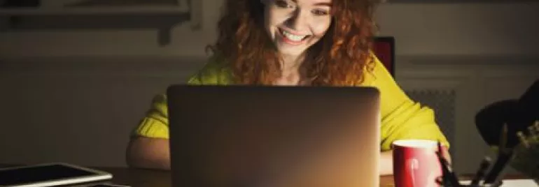 Woman with red hair smiling at a laptop screen in a dimly lit room.