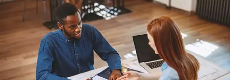 Two people discussing over documents at a table with laptops in a bright office.
