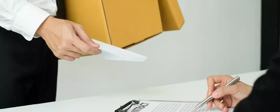 Person handing an envelope to someone signing a contract at a desk.