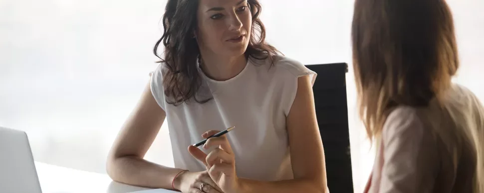Two women having a conversation at a desk with a laptop nearby.