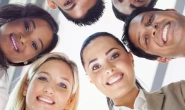 Group of six diverse people smiling, standing in a circle looking down.