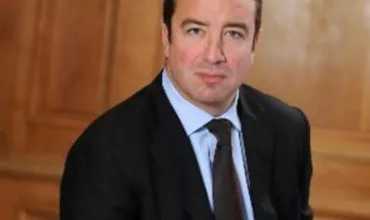 Man in a suit sitting with a wooden panel background.