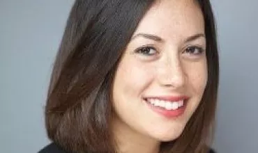 Portrait of a woman with shoulder-length dark hair, wearing a black blazer, smiling against a gray background.