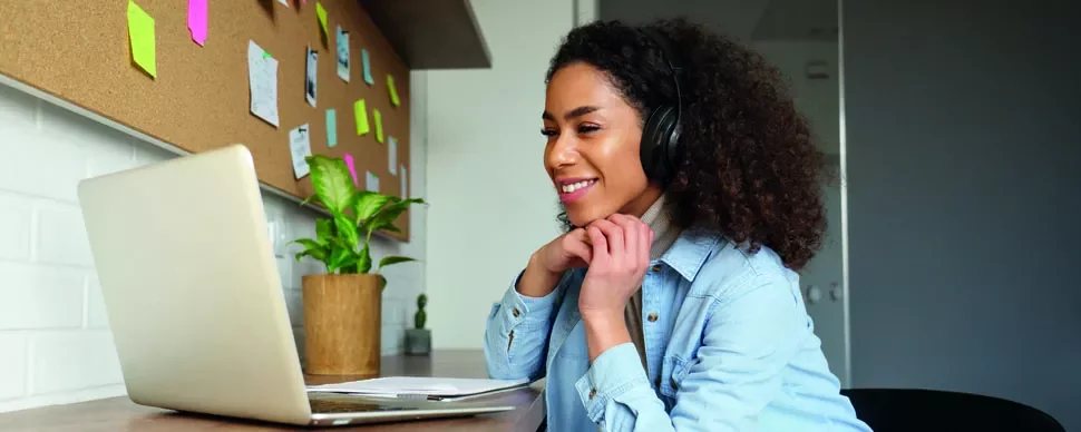 Woman with headphones smiling at laptop, working at home office desk.