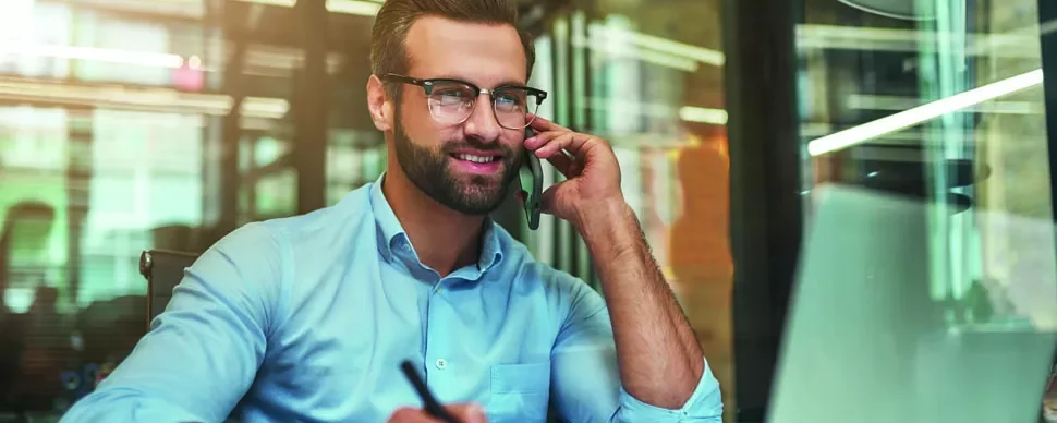 Man in a blue shirt talking on a phone and using a laptop at an office desk.