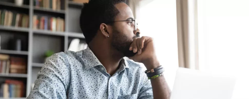 Man in patterned shirt looking thoughtfully out a window near a laptop.