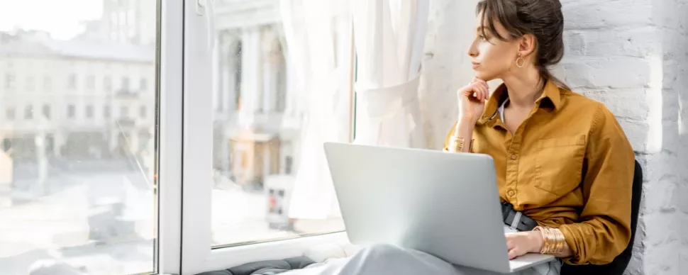 Woman sitting by a window with a laptop, looking outside thoughtfully.