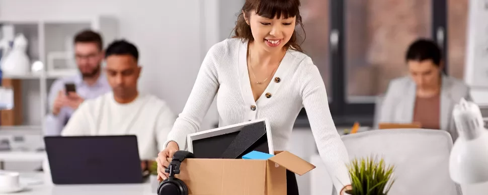 Woman unpacking office items from a box, smiling. Two colleagues work in the background.
