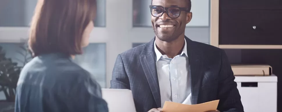 Man in glasses smiling while holding papers in an office meeting with a woman.