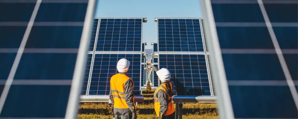 Two workers in hard hats and vests inspect large solar panels in a field.