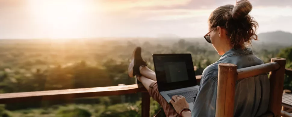 Person using a laptop on a balcony, overlooking a scenic sunset.