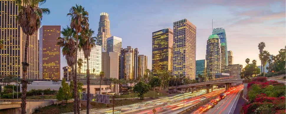 Los Angeles skyline at dusk with illuminated buildings and palm trees.