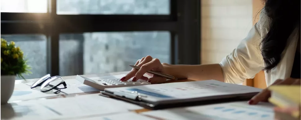 Person using a calculator at a desk with reports and glasses.