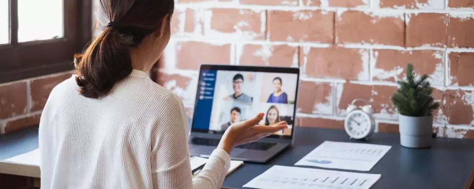 Person in a white sweater attending a video conference on a laptop at a desk with papers and a small plant.