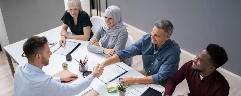 A diverse group of people having a meeting around a table, with two men shaking hands.