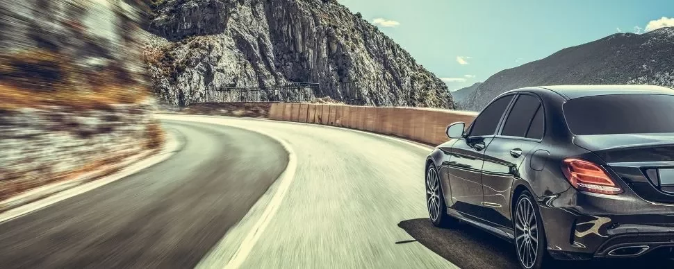 A sleek black car driving on a winding mountain road under a clear sky.