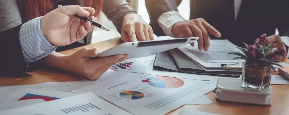 Hands holding a tablet over a table with charts and graphs.