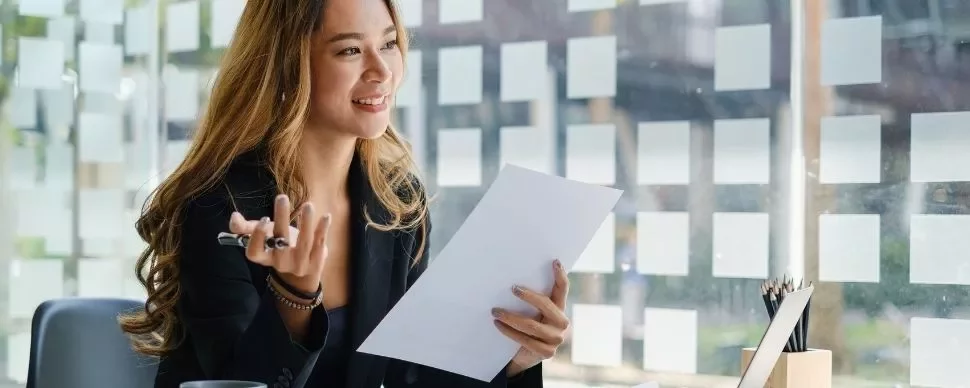 Smiling woman in an office holding a document, seated by a window with sticky notes.