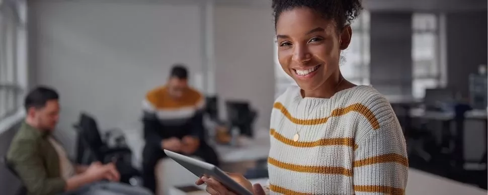 Woman in striped sweater smiling, holding a tablet in office.