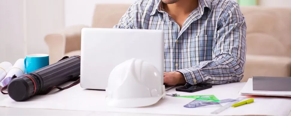 Man in plaid shirt working on a laptop with a white helmet on the table.