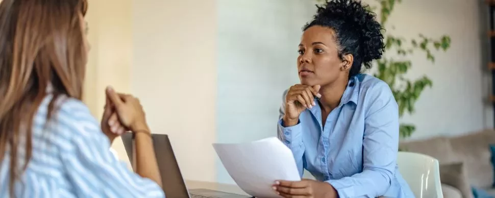 Two women having a focused discussion at a desk with a laptop and paperwork.