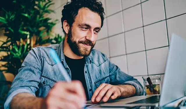 Man in denim shirt smiling while working on a laptop at a desk.